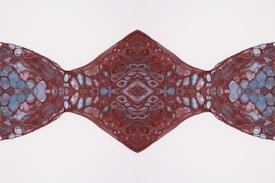 An abstract design featuring symmetrical patterns resembling organic or cellular structures. The patterns are predominantly in shades of red, with accents of blue and white, creating a kaleidoscopic effect.