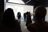 Silhouettes of people in a dimly lit room are captured from behind, facing a large display. The display showcases elegant suits and clothing. One person is holding a phone, taking a picture of the scene, with a clock face seen through a window in the background.