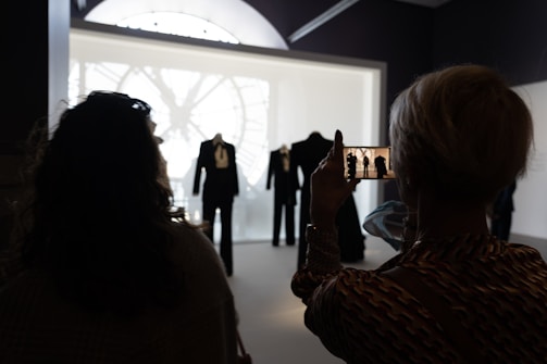 Silhouettes of people in a dimly lit room are captured from behind, facing a large display. The display showcases elegant suits and clothing. One person is holding a phone, taking a picture of the scene, with a clock face seen through a window in the background.