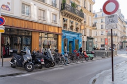 A city street scene featuring a row of parked motorcycles and bicycles in front of a clothing store with large windows displaying various garments. The buildings are classic European architecture with balconies and window boxes containing flowers. A street sign is visible in the foreground, and pedestrians can be seen walking along the sidewalk.