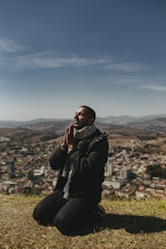 A person kneels on a grassy hilltop with hands clasped in prayer, overlooking a sprawling cityscape below. The clear blue sky stretches above with distant mountains in the background.