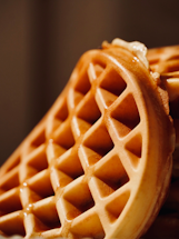 Golden brown waffles stacked at an angle, with visible syrup glistening on the surface highlighting the texture and grid pattern.