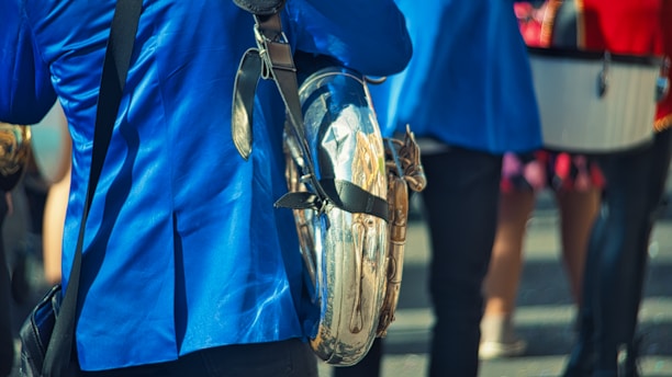 Several individuals wearing bright blue jackets and holding musical instruments are gathered in a group. A shiny sousaphone is prominently displayed, reflecting light and some surrounding elements. The scene appears to be outdoors with a casual, lively atmosphere.
