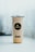A clear cup filled with a light-colored beverage, featuring a black and gold logo with the word 'Majestea' on the front. The cup is sealed with a silver foil lid and has social media icons displayed below the logo. The background is softly blurred, creating a focus on the cup.