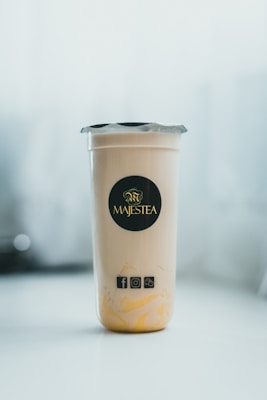 A clear cup filled with a light-colored beverage, featuring a black and gold logo with the word 'Majestea' on the front. The cup is sealed with a silver foil lid and has social media icons displayed below the logo. The background is softly blurred, creating a focus on the cup.