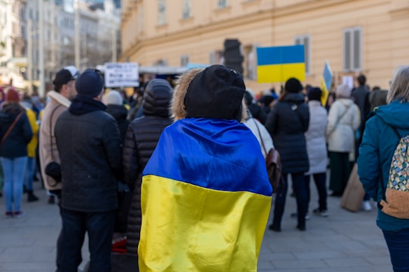A group of people gathered, some holding signs and flags, with one person prominently wearing a Ukrainian flag draped over their shoulders. The setting appears to be urban, with buildings and a street visible in the background. The crowd is dressed in winter clothing, suggesting a cold day.