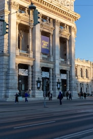 A grand, historical building with tall columns and ornate architectural details stands prominently. A large banner with the text '#Stand With Ukraine' hangs between the columns. Several people walk along the sidewalk in front of the building, casting long shadows in the early evening light.