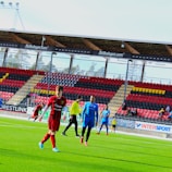 A group of soccer players are on a green pitch in a stadium, some wearing red and black uniforms while others are in blue. The stadium has empty stands with red, black, and yellow seats and advertisements. Trees are visible through the transparent stadium roof.