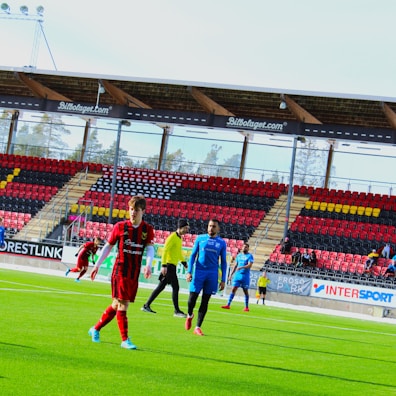 A group of soccer players are on a green pitch in a stadium, some wearing red and black uniforms while others are in blue. The stadium has empty stands with red, black, and yellow seats and advertisements. Trees are visible through the transparent stadium roof.