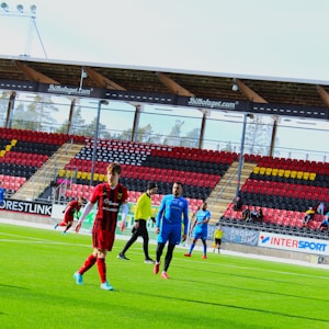 A group of soccer players are on a green pitch in a stadium, some wearing red and black uniforms while others are in blue. The stadium has empty stands with red, black, and yellow seats and advertisements. Trees are visible through the transparent stadium roof.
