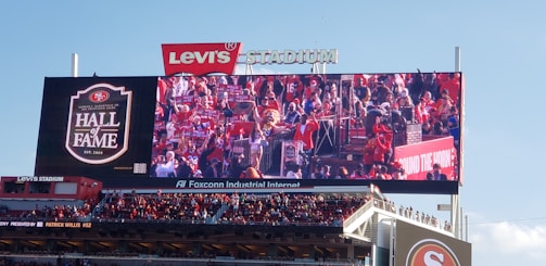 A large crowd is gathered at an outdoor stadium, where a big electronic screen displays an image of the audience members celebrating passionately. The sign 'Hall of Fame' is visible on the screen along with logos and branding. The sky is clear and blue.