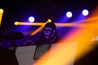 A DJ wearing a black hood and a white skull mask is performing on stage. Bright yellow and blue lights illuminate the scene, creating a dramatic and vibrant atmosphere. Light beams cross each other, adding dynamic energy to the environment.
