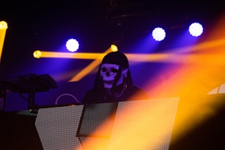 A DJ wearing a black hood and a white skull mask is performing on stage. Bright yellow and blue lights illuminate the scene, creating a dramatic and vibrant atmosphere. Light beams cross each other, adding dynamic energy to the environment.