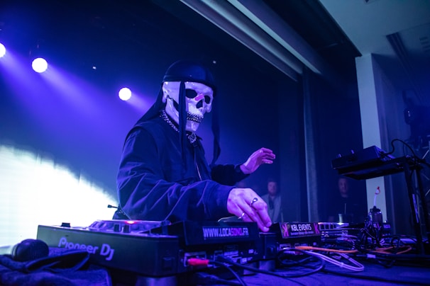 A DJ wearing a skull mask and dark clothing is performing at a club or concert venue. The artist is surrounded by DJ equipment, including mixers and other electronic music devices. The background is filled with purple and blue stage lights, creating a vibrant atmosphere. There are a few people in the audience, partially visible in the dimly lit setting.