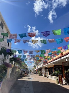 Colorful papel picado banners are suspended across a lively pedestrian street. The scene is bustling with vibrant energy, flanked by shops on either side and lush green plants. A clear blue sky with scattered clouds adds brightness to the scene, enhancing the festive atmosphere.