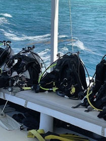 Several scuba diving gear sets, including tanks and hoses, are neatly arranged on a bench on a boat. The background shows clear blue ocean water with gentle waves, indicating a serene environment perfect for diving activities.