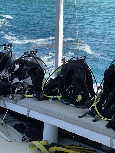Several scuba diving gear sets, including tanks and hoses, are neatly arranged on a bench on a boat. The background shows clear blue ocean water with gentle waves, indicating a serene environment perfect for diving activities.