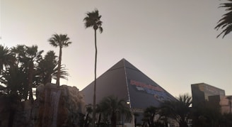 A modern pyramid-shaped building is surrounded by palm trees and a man-made waterfall. The sky is clear, suggesting it might be early evening or late afternoon. The structure has signage indicating an entertainment event.