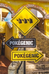 A vibrant street sign depicting silhouettes of a popular fictional character's head in profile, in a line on a yellow diamond-shaped sign. Below, black and yellow directional signs with the word 'Pokégenic' point in opposite directions against a background of stone steps and a decorative yellow building.