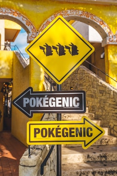 A vibrant street sign depicting silhouettes of a popular fictional character's head in profile, in a line on a yellow diamond-shaped sign. Below, black and yellow directional signs with the word 'Pokégenic' point in opposite directions against a background of stone steps and a decorative yellow building.