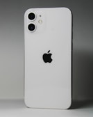 A white smartphone featuring a dual-camera setup in the upper left corner and a prominent black apple logo in the center. The device is positioned against a gradient gray background.