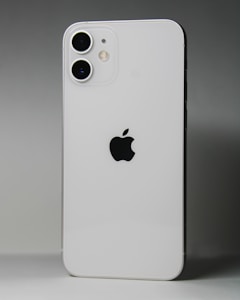 A white smartphone featuring a dual-camera setup in the upper left corner and a prominent black apple logo in the center. The device is positioned against a gradient gray background.