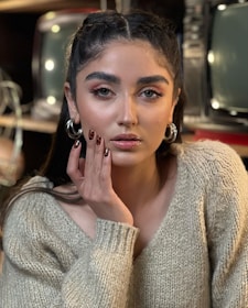 A person with long dark hair and hoop earrings gazes directly at the camera with a neutral expression. The individual is wearing a beige knitted sweater and has their hand gently touching their cheek, displaying manicured nails with dark polish. Old television sets are in the background, slightly out of focus.