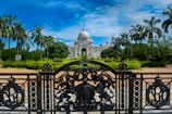 An ornate iron gate in the foreground leads to a grand white marble building with a large dome and multiple smaller domes, surrounded by well-maintained gardens with tall palm trees. The sky above is bright blue with some clouds.