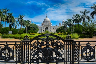 An ornate iron gate in the foreground leads to a grand white marble building with a large dome and multiple smaller domes, surrounded by well-maintained gardens with tall palm trees. The sky above is bright blue with some clouds.