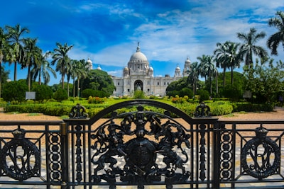 An ornate iron gate in the foreground leads to a grand white marble building with a large dome and multiple smaller domes, surrounded by well-maintained gardens with tall palm trees. The sky above is bright blue with some clouds.