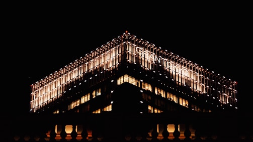 A brightly lit modern building with a prominent grid-like facade stands against a dark night sky. The illumination highlights the structure's architectural details and the text 'GRANTOTEL' is visible on one side of the top.