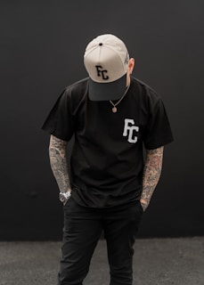 A person is standing with their head slightly bowed, wearing a black t-shirt with 'FC' on it and a two-tone cap featuring the same letters. Their arms are adorned with tattoos, and they are wearing a watch on one wrist.