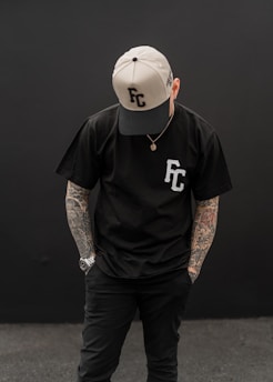 A person is standing with their head slightly bowed, wearing a black t-shirt with 'FC' on it and a two-tone cap featuring the same letters. Their arms are adorned with tattoos, and they are wearing a watch on one wrist.