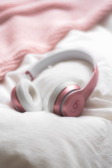 A pair of pink over-ear headphones is resting on a cozy, white fabric surface. In the background, there is a soft, pink blanket.