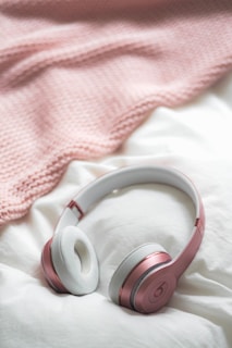 A pair of stylish over-ear headphones with a rose gold metallic finish and white cushioning is placed on a soft, white fabric. In the background, there's a textured light pink blanket, adding a cozy touch.
