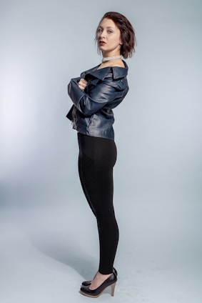 A person stands confidently in a profile pose with arms crossed, wearing a stylish leather jacket and black leggings. The individual has short, dark hair and is accessorized with a choker necklace and black high heels. The background is a plain, neutral shade that accentuates the subject.