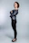 A person stands confidently in a profile pose with arms crossed, wearing a stylish leather jacket and black leggings. The individual has short, dark hair and is accessorized with a choker necklace and black high heels. The background is a plain, neutral shade that accentuates the subject.