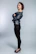 A person stands confidently in a profile pose with arms crossed, wearing a stylish leather jacket and black leggings. The individual has short, dark hair and is accessorized with a choker necklace and black high heels. The background is a plain, neutral shade that accentuates the subject.