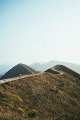 A scenic mountain path gently winds its way over rolling hills under a clear blue sky. The landscape is lush with green vegetation, and a lone hiker is seen walking along the trail, enjoying the tranquility and natural beauty of the surroundings. The distant mountains add depth to the background.