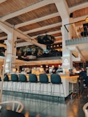 A modern interior of a spacious bar with high ceilings and wooden beams. The bar counter is illuminated with warm lighting, surrounded by high black chairs. Hanging plants are suspended from the ceiling, and there are people interacting near the bar. The ambiance is warm and inviting, with a mix of contemporary and rustic design elements.