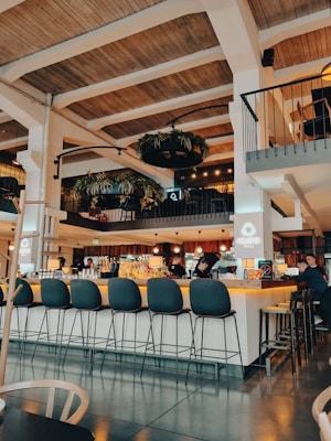 A modern interior of a spacious bar with high ceilings and wooden beams. The bar counter is illuminated with warm lighting, surrounded by high black chairs. Hanging plants are suspended from the ceiling, and there are people interacting near the bar. The ambiance is warm and inviting, with a mix of contemporary and rustic design elements.