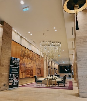 An elegantly designed hotel lobby featuring a large, artistic mural on the wall. The room is furnished with plush chairs around a central glass table on a patterned carpet. A prominent chandelier with hanging glass pieces is suspended from the high ceiling, and a stylish promotional stand is placed near one wall. The lighting is warm, creating an inviting atmosphere.
