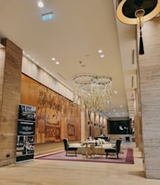 An elegantly designed hotel lobby featuring a large, artistic mural on the wall. The room is furnished with plush chairs around a central glass table on a patterned carpet. A prominent chandelier with hanging glass pieces is suspended from the high ceiling, and a stylish promotional stand is placed near one wall. The lighting is warm, creating an inviting atmosphere.