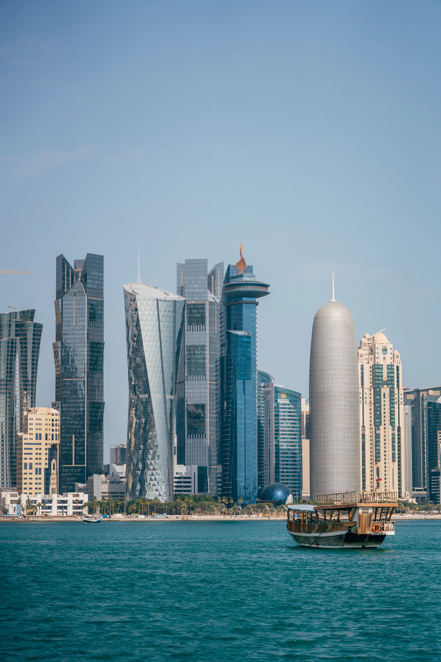 A wooden dhow crossing blue water in front of Doha's West Bay skyline