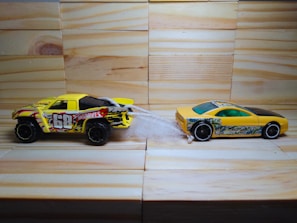 Two toy cars are positioned on a wooden surface with a light-colored wood paneling background. The car on the left is a yellow and black truck with flames and the number 68 on its side. It is connected to a yellow and black sports car on the right by a piece of white string or material.