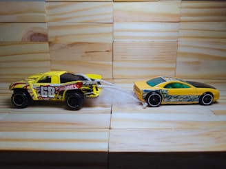 Two toy cars are positioned on a wooden surface with a light-colored wood paneling background. The car on the left is a yellow and black truck with flames and the number 68 on its side. It is connected to a yellow and black sports car on the right by a piece of white string or material.