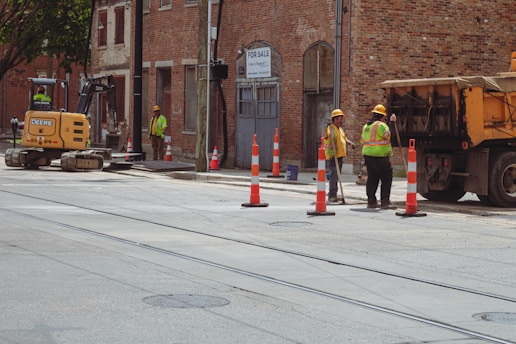 Construction workers wearing safety gear are at a street worksite. They are using heavy machinery like a small excavator and a dump truck. Traffic cones are placed around them, and a 'For Sale' sign is visible on a nearby building.