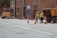 Construction workers wearing safety gear are at a street worksite. They are using heavy machinery like a small excavator and a dump truck. Traffic cones are placed around them, and a 'For Sale' sign is visible on a nearby building.