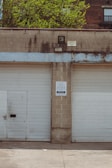 Two closed garage doors, separated by a concrete column, with a small door on the left. A sign with a warning is posted on the column. The building appears weathered with visible stains, and there are green trees and brick buildings in the background.