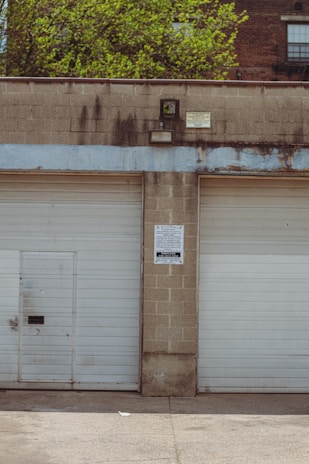 Two closed garage doors, separated by a concrete column, with a small door on the left. A sign with a warning is posted on the column. The building appears weathered with visible stains, and there are green trees and brick buildings in the background.